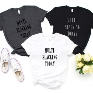 Puede incluir: Tres camisetas con el texto "MULTI-SLACKING TODAY" impreso en ellas. Las camisetas son de color gris, negro y blanco.