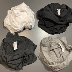 May include: Four folded t-shirts in white, black, charcoal gray, and light gray. Each shirt has a tag visible. The shirts are made of a soft material and are neatly folded, ready for wear.