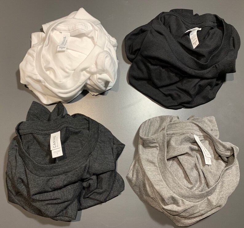 May include: Four folded t-shirts in white, black, charcoal gray, and light gray. Each shirt has a tag visible. The shirts are made of a soft material and are neatly folded, ready for wear.