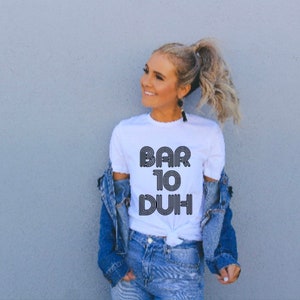 May include: White t-shirt with the text "BAR 10 DUH" in a retro font. The shirt is paired with a denim jacket and jeans. The overall style is casual and modern.