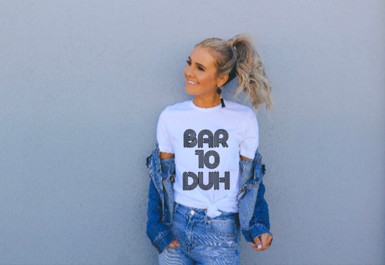 May include: White t-shirt with the text "BAR 10 DUH" in a retro font. The shirt is paired with a denim jacket and jeans. The overall style is casual and modern.