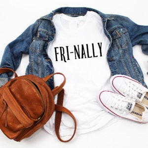 May include: White t-shirt with the text "FRI-NALLY" in black lettering. The t-shirt is paired with a denim jacket, a brown leather backpack, and white sneakers.