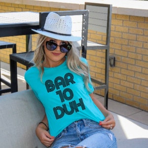 May include: A turquoise t-shirt with the text "BAR TO DUH" in black, worn with denim shorts and a white hat. The image is taken outdoors, with a brick wall and outdoor furniture in the background.
