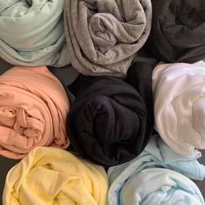 May include: An assortment of rolled-up t-shirts in various colors, including light green, gray, black, peach, yellow, and light blue. The shirts are neatly folded, showcasing the different hues and textures of the fabric. The arrangement suggests a focus on color variety and style.
