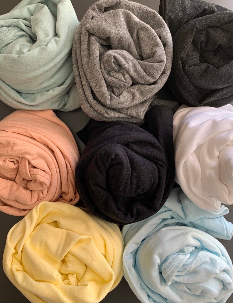 May include: An assortment of rolled-up t-shirts in various colors, including light green, gray, black, peach, yellow, and light blue. The shirts are neatly folded, showcasing the different hues and textures of the fabric. The arrangement suggests a focus on color variety and style.