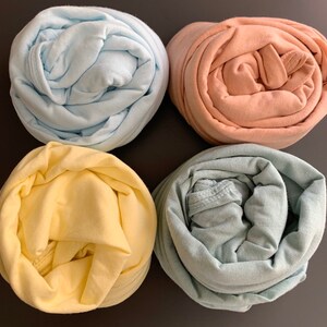 May include: Four rolled-up fabric items in pastel colors: light blue, peach, yellow, and a muted green. The fabric appears to be soft and possibly cotton or a similar material. The items are arranged on a dark surface.