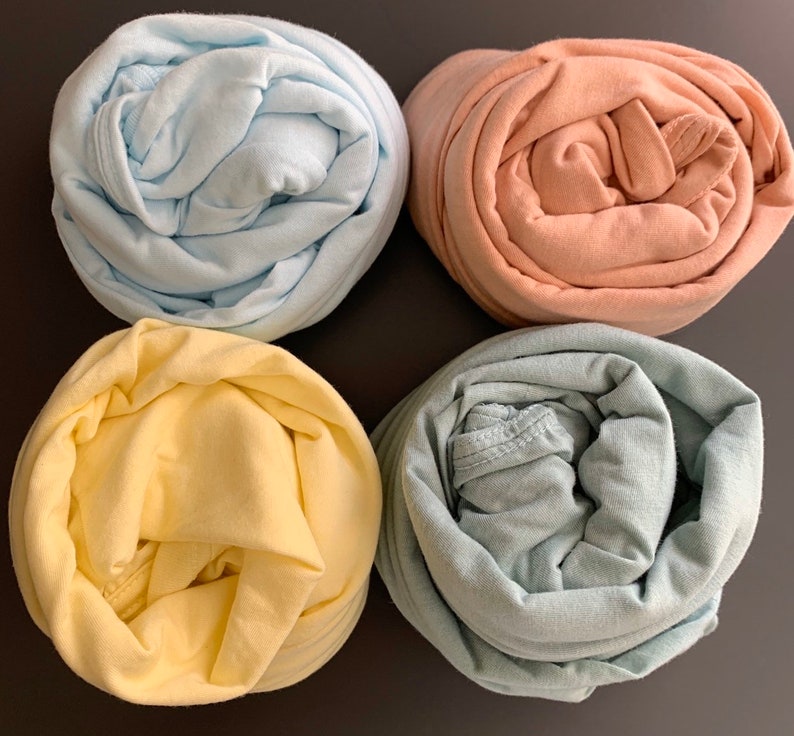 May include: Four rolled-up fabric items in pastel colors: light blue, peach, yellow, and a muted green. The fabric appears to be soft and possibly cotton or a similar material. The items are arranged on a dark surface.
