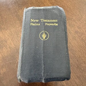 May include: A black, textured, hardbound book with gold lettering on the front cover that reads New Testament, Psalms, and Proverbs. Below the text is a small, circular gold emblem featuring an illustration of an oil lamp or vessel.
