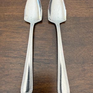 Puede incluir: Dos cucharas de pomelo de acero inoxidable con bordes dentados y mangos largos con cuentas. Las cucharas se exhiben sobre una superficie de madera oscura, mostrando su acabado pulido y diseño.