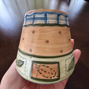 May include: A ceramic mug featuring a tan upper section with brown polka dots, a blue and white plaid rim, and a light green lower band decorated with rectangular panels containing dark green leaf illustrations and stitched border details.