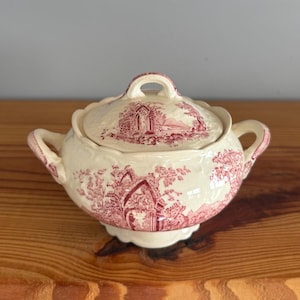 May include: A cream-colored ceramic sugar bowl with a matching lid, featuring a red transferware pattern of architectural ruins and trees. The bowl has two side handles, a scalloped rim, and a pedestal base with a matching red landscape design.