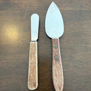 May include: Two cheese knives with wooden handles. One is a small butter knife, and the other is a larger, flat cheese knife. The blades are silver, and the handles are a light brown color.