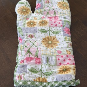 May include: A quilted oven mitt featuring a patchwork design with floral and butterfly motifs. The mitt showcases a mix of pastel colors, including pink, yellow, and green, with a green gingham trim. The mitt is designed for kitchen use.