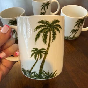 May include: White ceramic mugs with a palm tree design. The mugs have a curved handle and a cylindrical shape. The palm tree design features green fronds and a brown trunk. The mugs are set on a dark brown wooden surface.