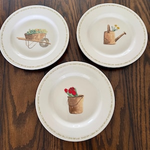 May include: Three off-white ceramic plates featuring garden-themed illustrations. One plate shows a wheelbarrow with flowers, another a watering can with yellow flowers, and the third a bucket with red tulips. Each rim bears the text NATURE GIVES US THE VARIETY WE CRAVE.