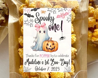Può includere: Un sacchetto di plastica trasparente pieno di popcorn gialli con un'etichetta bianca e rosa che dice "Spooky one!" con un'illustrazione di un fantasma e una zucca. L'etichetta dice anche "Thanks for POPPIN' by to celebrate Madeline's 1st Boo-Day! October 7, 2023."