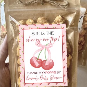 May include: Clear cellophane bag filled with popcorn, tied with twine. A pink label features a cherry illustration and the text "SHE IS THE cherry on top! THANKS FOR POPPIN' BY Emma's Baby Shower."