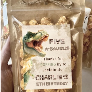 May include: Clear cellophane bag filled with caramel popcorn, tied with twine. A label features a green and tan dinosaur illustration and the text "FIVE A-SAURUS" and "CHARLIE'S 5TH BIRTHDAY".