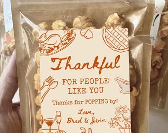 Puede incluir: Una bolsa de plástico transparente llena de palomitas de maíz, sellada con una parte superior de papel marrón y un lazo de cuerda de yute. Una etiqueta rectangular presenta dibujos lineales naranjas de un pavo, pastel, calabaza y flores con el texto: Thankful FOR PEOPLE LIKE YOU Thanks for POPPING by! Love, Brad & Jenn.