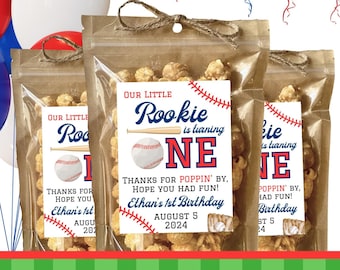 May include: A set of three brown paper bags filled with popcorn and tied with twine. Each bag has a white label with a baseball theme and the text "Our Little Rookie is turning One". The labels also include the text "Ethan's 1st Birthday" and the date "August 5, 2024".
