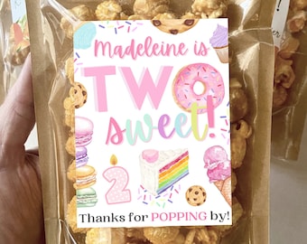 May include: A clear plastic bag filled with caramel popcorn with a pink and white tag that says "madeleine is TWO sweet!" with a rainbow cake, ice cream cone, donut, and macarons. The tag also says "Thanks for POPPING by!"
