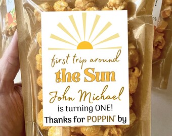 May include: A clear plastic bag filled with caramel popcorn with a yellow and white sun graphic on a white tag. The tag reads "first trip around the Sun John Michael is turning ONE! Thanks for POPPIN' by"