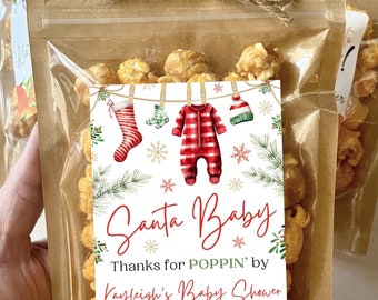 Op de afbeelding: Een kleine doorzichtige plastic zak gevuld met popcorn en vastgebonden met touw. Een wit label met een rode en groene illustratie van een baby in een kerstmanpak dat aan een waslijn hangt met sneeuwvlokken en groen. Het label luidt "Santa Baby Thanks for POPPIN' by Kayleigh's Baby Shower December 9, 2023".