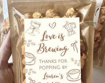 Peut inclure: Un sachet en plastique transparent rempli de pop-corn couleur caramel, scellé avec un haut en papier brun et de la ficelle. Une étiquette blanche présente des dessins au trait brun d'articles de café et le texte : Love is Brewing, THANKS FOR POPPING BY, Lauren's Bridal Shower.