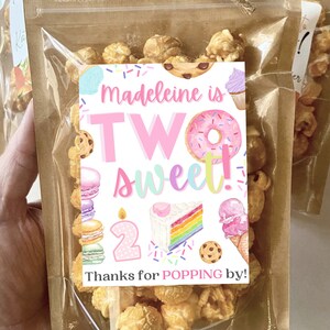 May include: A clear plastic bag filled with caramel popcorn with a pink and white tag that says "madeleine is TWO sweet!" with a rainbow cake, ice cream cone, donut, and macarons. The tag also says "Thanks for POPPING by!"