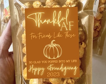 May include: A brown paper bag filled with caramel popcorn with a label that says "Thankful AF For Friends Like These" with a pumpkin graphic and the text "So glad you popped into my life! Happy Friendsgiving 2023".