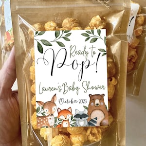 May include: A clear plastic pouch filled with caramel popcorn, sealed with a brown paper top and a jute twine bow. A white label features green leaves, the text Ready to Pop! Lauren's Baby Shower October 2023, and illustrations of a deer, fox, raccoon, and bear.