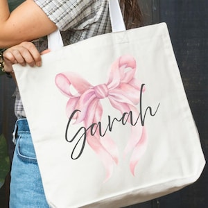 May include: A cream-coloured tote bag featuring a pink watercolour bow and the name "Sarah" in cursive writing. The bag has white handles and is held by a person wearing sunglasses and a plaid shirt.