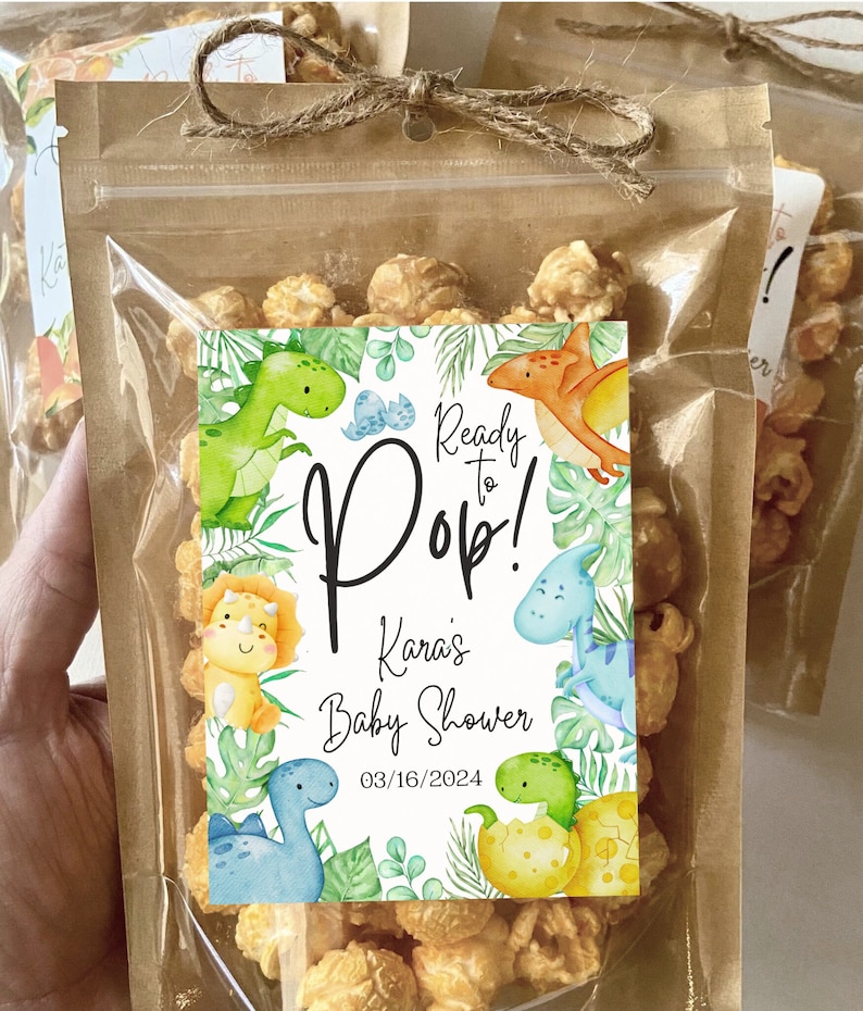 May include: A brown paper bag with a clear plastic window filled with caramel popcorn. The bag is tied with a brown twine bow. A white sticker with green and blue dinosaur illustrations is attached to the bag. The sticker reads 'Ready to Pop! Kara's Baby Shower 03/16/2024'.