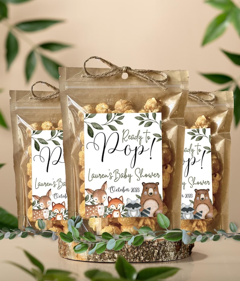 May include: Three clear plastic pouches filled with popcorn, each featuring a label with the text Ready to Pop! Lauren's Baby Shower October 2023. The labels display watercolor illustrations of a deer, fox, raccoon, and bear with green leaves.