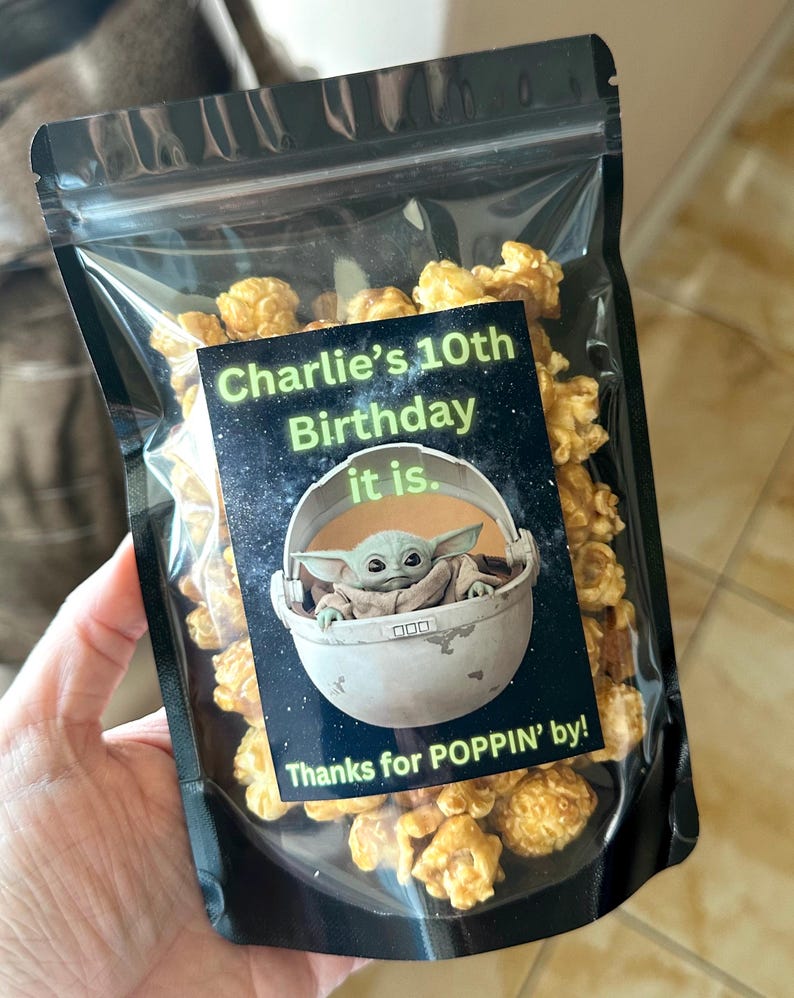 May include: A clear plastic bag filled with caramel popcorn. A black and green sticker with a picture of a character from Star Wars, Baby Yoda, is on the bag. The sticker says "Charlie's 10th Birthday it is. Thanks for POPPIN' by!"