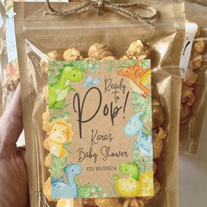 May include: A clear plastic bag filled with popcorn and a brown paper tag with a dinosaur theme. The tag reads "Ready to Pop! Kara's Baby Shower 03/16/2024".