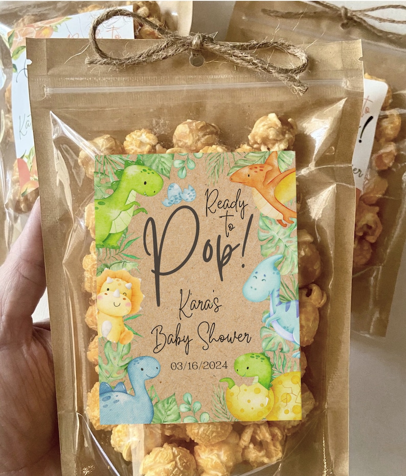 May include: A clear plastic bag filled with popcorn and a brown paper tag with a dinosaur theme. The tag reads "Ready to Pop! Kara's Baby Shower 03/16/2024".