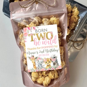 May include: A pink and white favor bag filled with popcorn and a pink and gold tag that says "Born Two Be Wild" with a floral design and a giraffe, elephant, lion, and zebra. The tag also says "Thanks for popping by Annie's 2nd Birthday! 07/21/24"