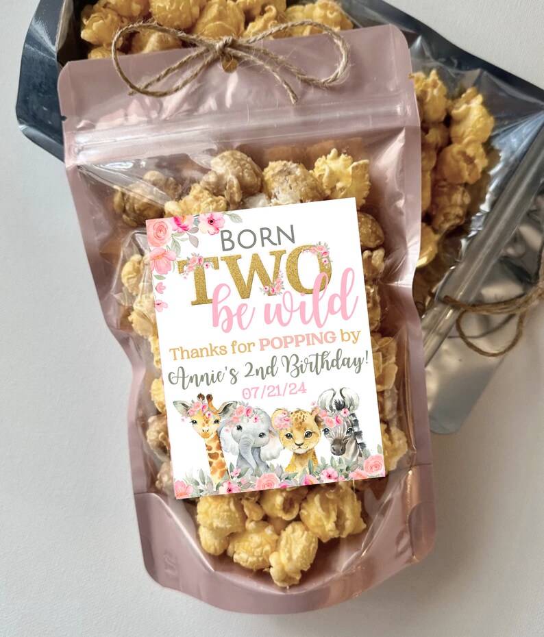 May include: A pink and white favor bag filled with popcorn and a pink and gold tag that says "Born Two Be Wild" with a floral design and a giraffe, elephant, lion, and zebra. The tag also says "Thanks for popping by Annie's 2nd Birthday! 07/21/24"