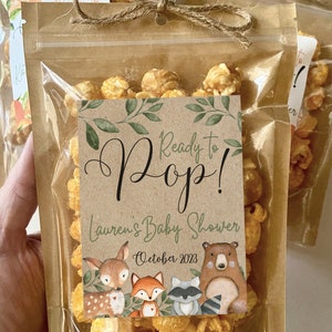 May include: A clear plastic pouch filled with golden caramel popcorn, sealed with a brown paper top and a jute twine bow. A rectangular label features the text Ready to Pop! Lauren's Baby Shower October 2023, decorated with green leaves and illustrations of a deer, fox, raccoon, and bear.