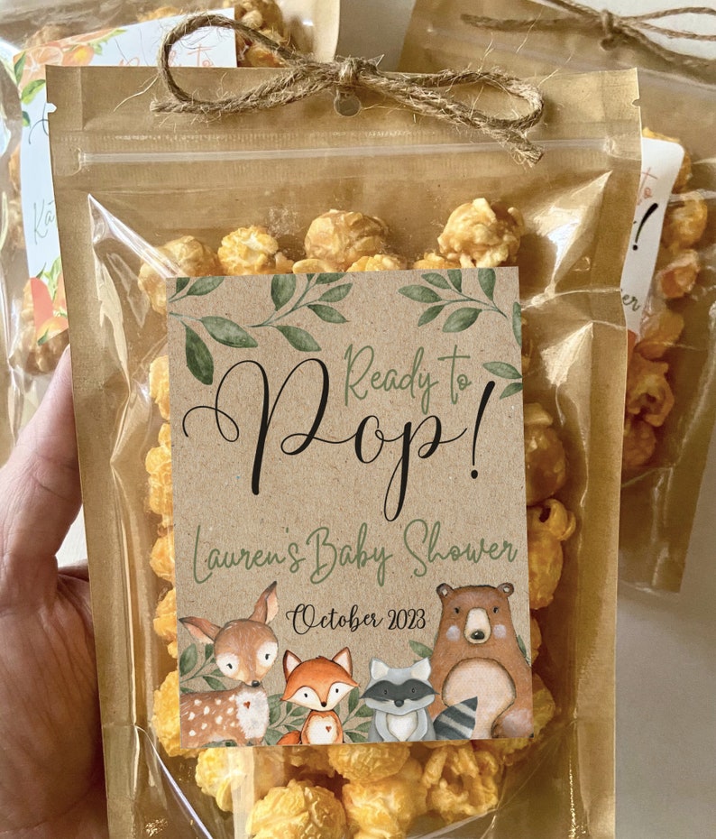 May include: A clear plastic pouch filled with golden caramel popcorn, sealed with a brown paper top and a jute twine bow. A rectangular label features the text Ready to Pop! Lauren's Baby Shower October 2023, decorated with green leaves and illustrations of a deer, fox, raccoon, and bear.