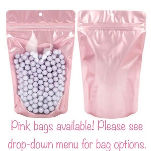 May include: Two pink, resealable bags. One bag is filled with small, round, light purple candies. The other bag is empty. The bags have a clear window to view the contents. The text "Pink bags available! Please see drop-down menu for bag options." is at the bottom.