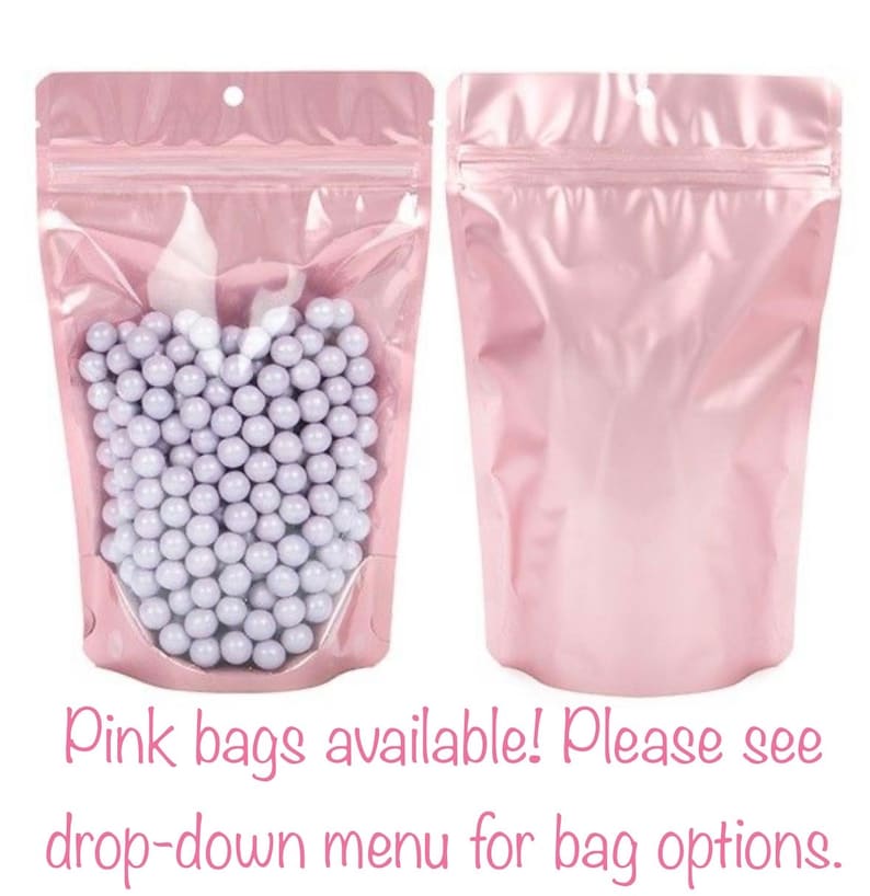 May include: Two pink, resealable bags. One bag is filled with small, round, light purple candies. The other bag is empty. The bags have a clear window to view the contents. The text "Pink bags available! Please see drop-down menu for bag options." is at the bottom.