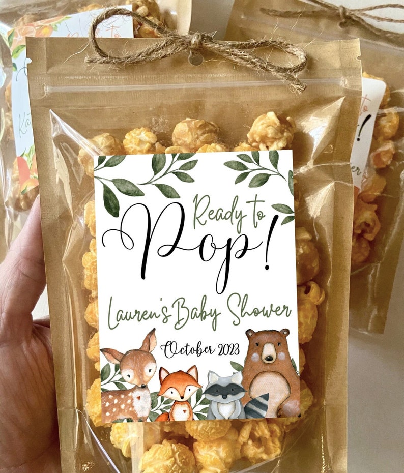 May include: A clear plastic pouch filled with caramel popcorn, sealed with a brown paper top and a jute twine bow. A white label features green leaves, the text Ready to Pop! Lauren's Baby Shower October 2023, and illustrations of a deer, fox, raccoon, and bear.