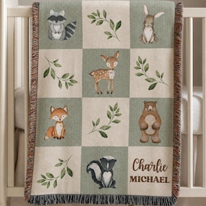 May include: A woven throw blanket featuring a grid pattern of alternating beige and sage green squares. The squares display illustrations of a raccoon, rabbit, deer, fox, bear, skunk, and green leaves. The name Charlie Michael is printed in brown.