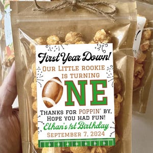 May include: A clear plastic pouch filled with caramel popcorn, tied at the top with brown twine. A white label features a football graphic, the text First Year Down! Our Little Rookie Is Turning One, Thanks For Poppin' By, Hope You Had Fun!, Ethan's 1st Birthday, September 7, 2024, and a green football field yard line graphic.