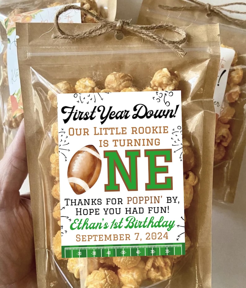 May include: A clear plastic pouch filled with caramel popcorn, tied at the top with brown twine. A white label features a football graphic, the text First Year Down! Our Little Rookie Is Turning One, Thanks For Poppin' By, Hope You Had Fun!, Ethan's 1st Birthday, September 7, 2024, and a green football field yard line graphic.