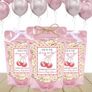 May include: Three pink cellophane bags filled with popcorn, each adorned with a label featuring red cherries and the text "She is the cherry on top!" and "Thanks for Poppin by Emma's Baby Shower". The bags are tied with a white ribbon or twine. Pink and silver balloons float in the background.