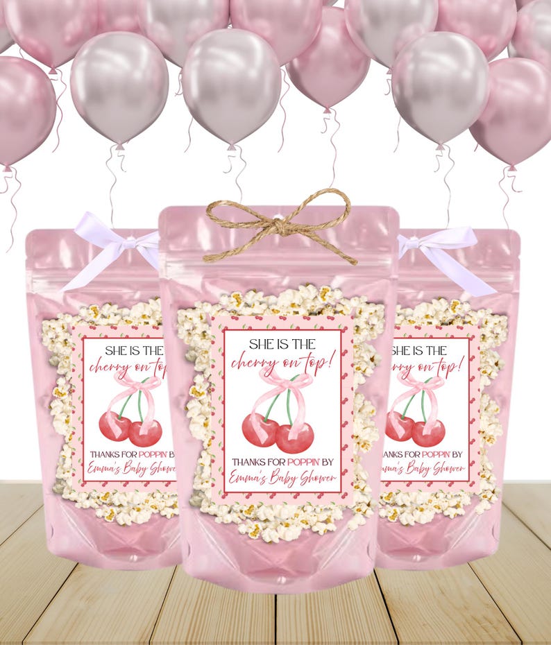 May include: Three pink cellophane bags filled with popcorn, each adorned with a label featuring red cherries and the text "She is the cherry on top!" and "Thanks for Poppin by Emma's Baby Shower". The bags are tied with a white ribbon or twine. Pink and silver balloons float in the background.