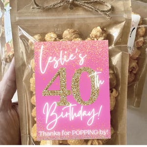 Pu&ograve; includere: Un sacchetto per bomboniere di compleanno rosa e oro pieno di popcorn al caramello. Il sacchetto ha un'etichetta che dice "Leslie's 40th Birthday! Thanks for POPPING by!"